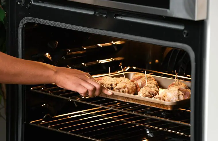 A person putting a cookie sheet of Sweet and Spicy Bacon Wrapped BBQ Chicken Strips into the oven