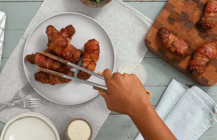 An overhead shot of a person placing finished Sweet and Spicy Bacon Wrapped BBQ Chicken Strips onto a plate using tongs